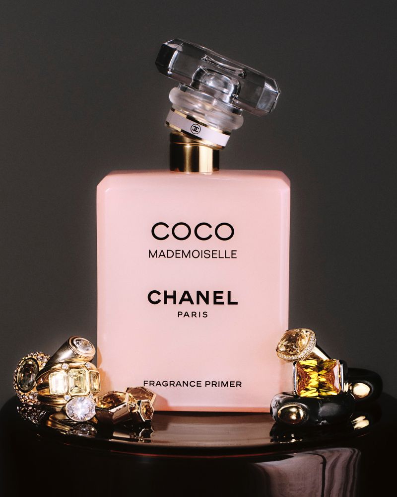 Still life studio shot of Coco Mademoiselle Fragrance Primer by Chanel, displayed with gold rings on a reflective black surface.