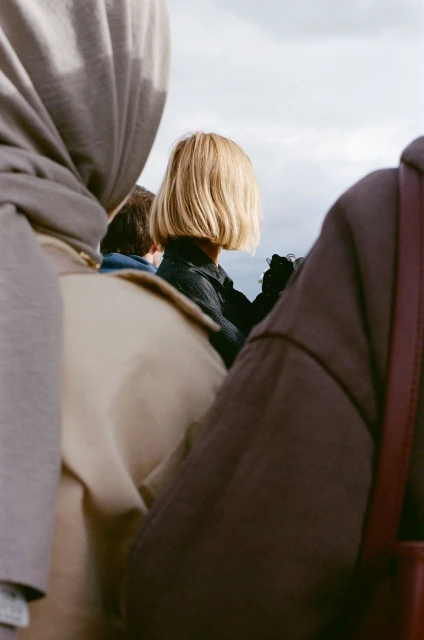 A group of people, including one with a blonde bob haircut, stand outside on a cloudy day, captured in a medium shot with natural lighting.