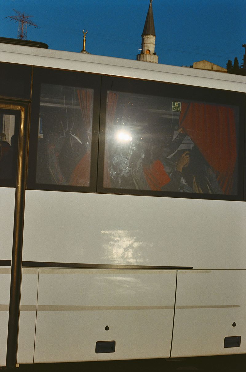 Exterior of a white bus with passengers visible through the window, against a blue sky with a minaret in the background.