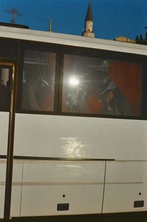 Exterior of a white bus with passengers visible through the window, against a blue sky with a minaret in the background.