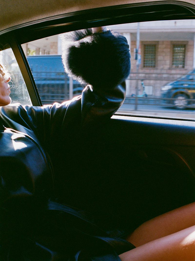 Young woman in the back seat of a car, looking out the window, wearing a black fur hat and jacket, with urban buildings in the background.