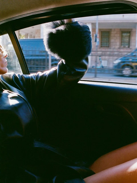 Young woman in the back seat of a car, looking out the window, wearing a black fur hat and jacket, with urban buildings in the background.