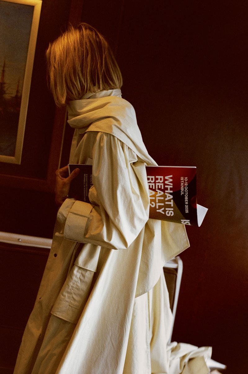 A blonde person wearing a cream-colored trench coat holding a brochure titled What is Really Real against a dark interior wall.
