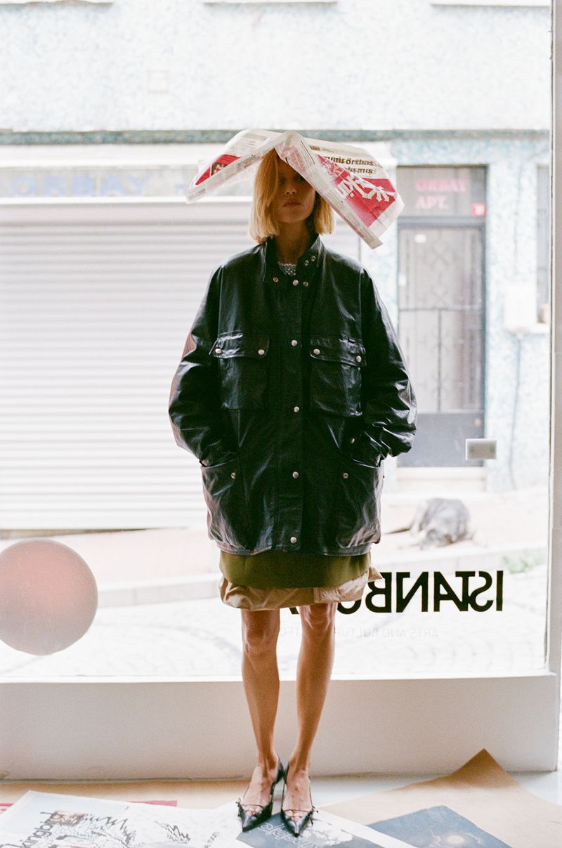 Full-length portrait of a blonde woman wearing a black jacket and newspaper hat, standing in front of a window with a reversed text.