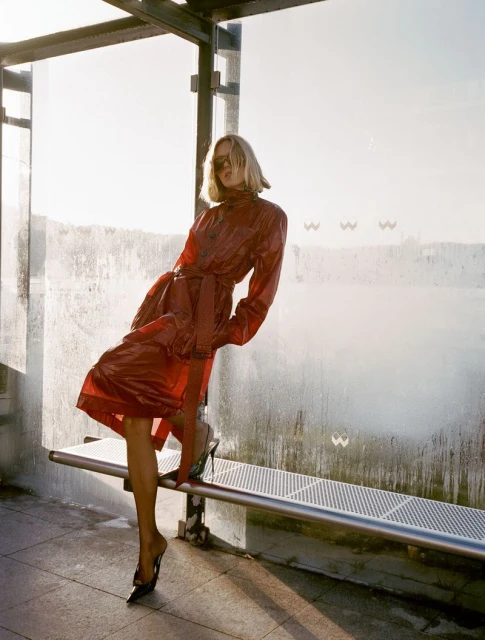 A blonde fashion model poses in a red vinyl coat and black high heels at an outdoor bus shelter with a bright, sunny background.