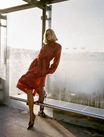 A blonde fashion model poses in a red vinyl coat and black high heels at an outdoor bus shelter with a bright, sunny background.