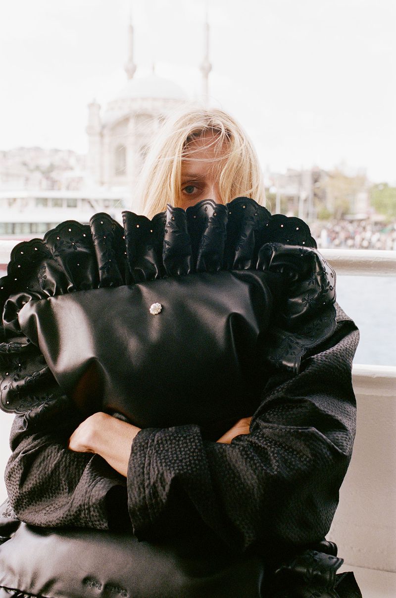 Woman holding a large black puffy bag in front of her face outdoors, wearing a black jacket with a Turkish mosque in the background.