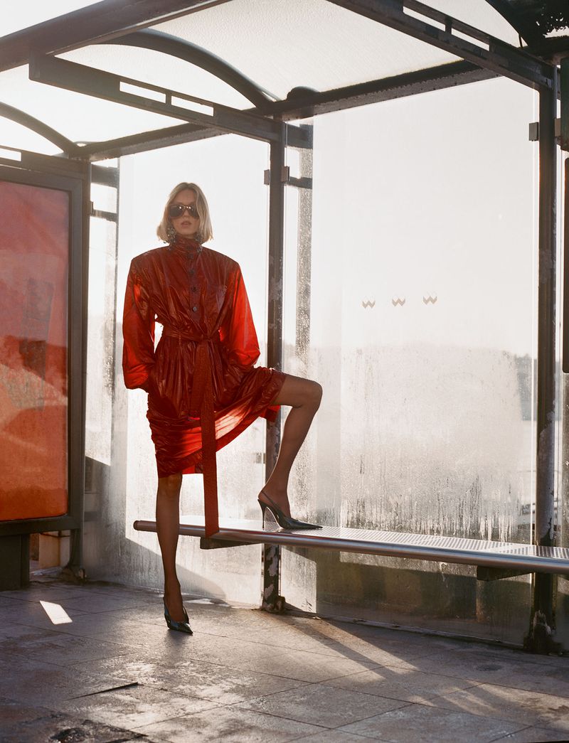 Blonde woman in a shiny red belted trench coat and dark sunglasses standing in a bus shelter against a bright sunlit background.