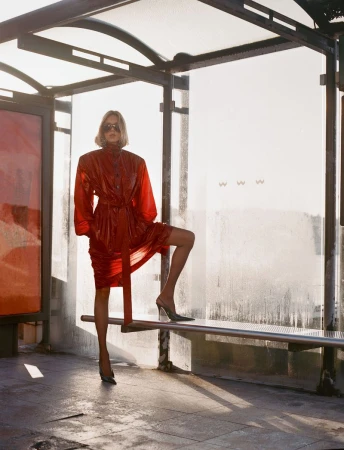 Blonde woman in a shiny red belted trench coat and dark sunglasses standing in a bus shelter against a bright sunlit background.