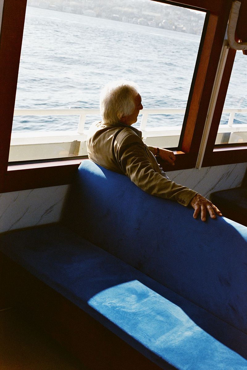 Elderly man gazes out a window aboard a boat, overlooking the blue water and distant shoreline on a bright day with sunlight.