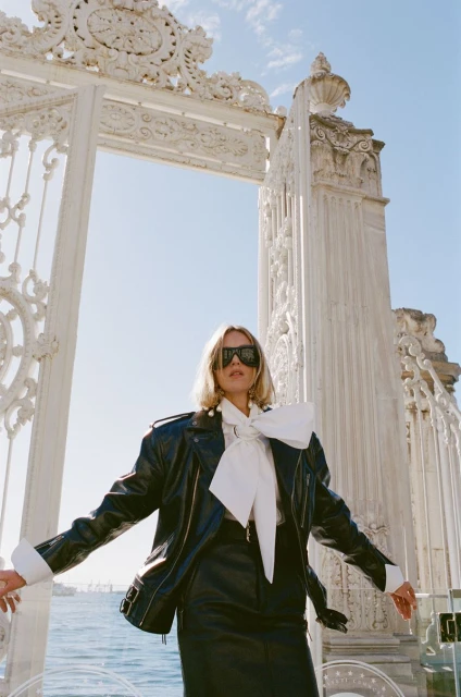 Blonde woman in a black leather jacket poses confidently against an ornate white archway and a blue sky background, fashion-forward style.