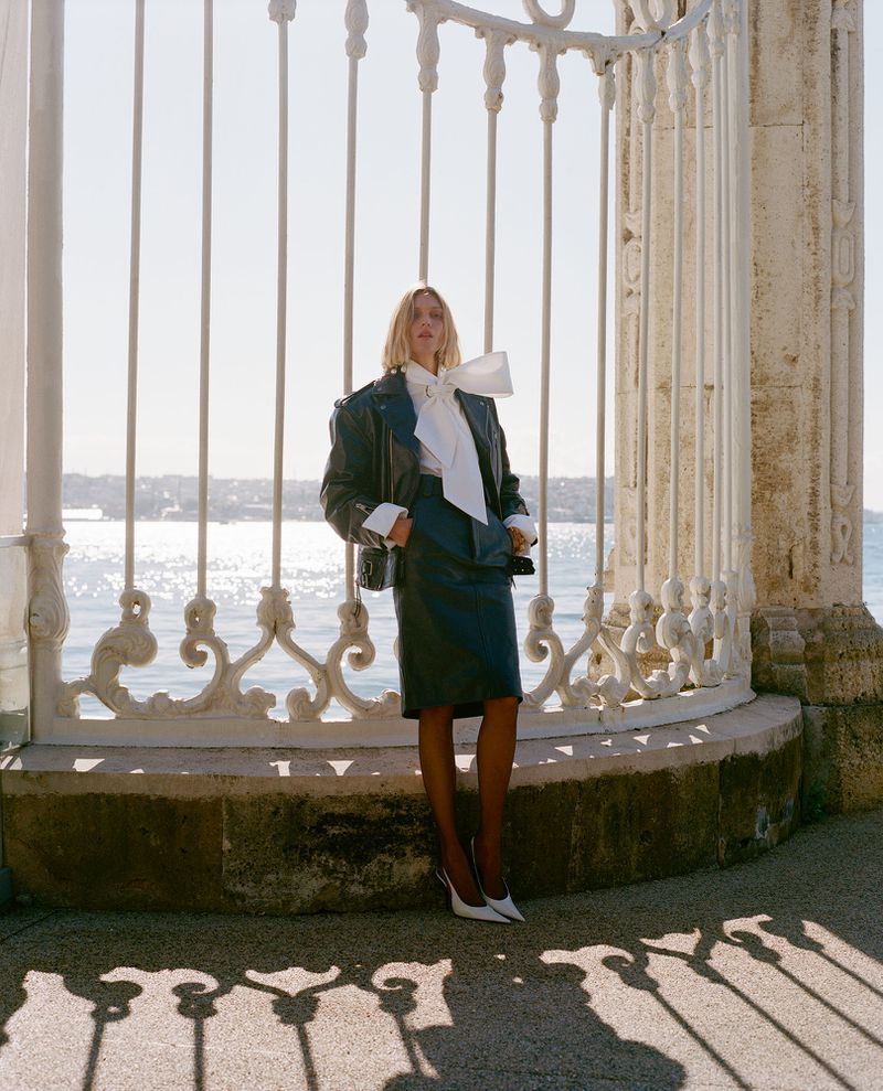 Woman in a black jacket and skirt poses near an ornate white fence with an ocean backdrop on a sunny day in an outdoor setting.