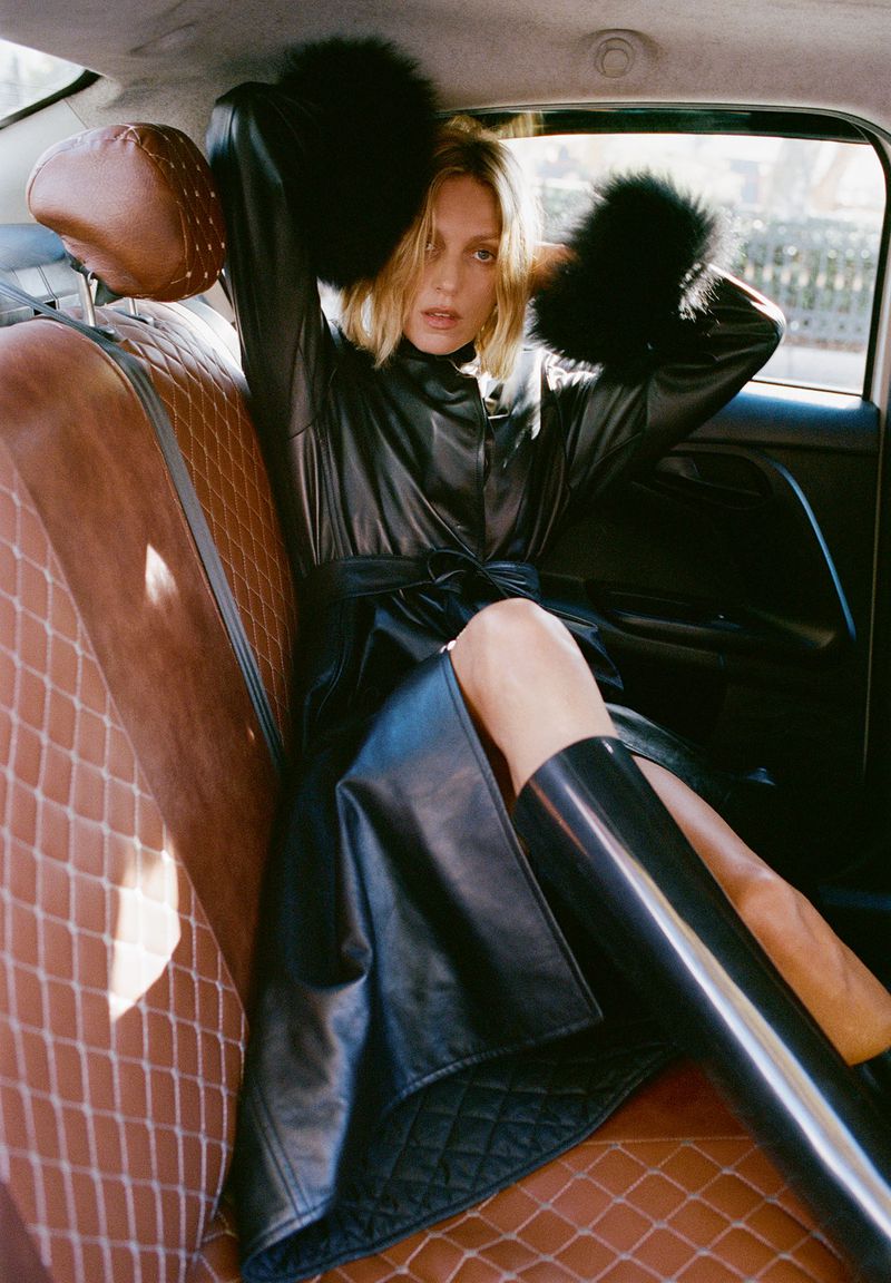 A blonde-haired woman in a black coat and boots sits inside of a car with tan quilted leather seats and black trim.
