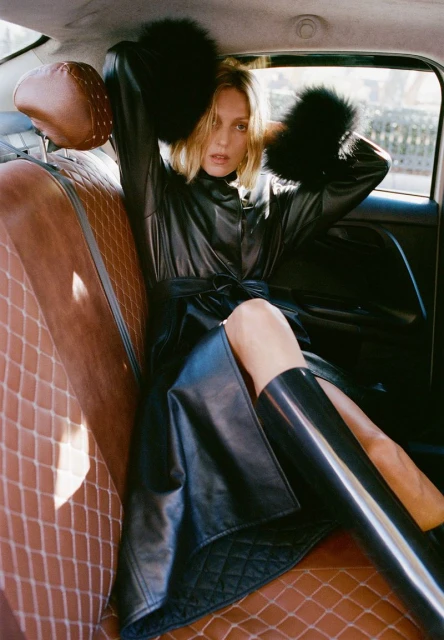 A blonde-haired woman in a black coat and boots sits inside of a car with tan quilted leather seats and black trim.