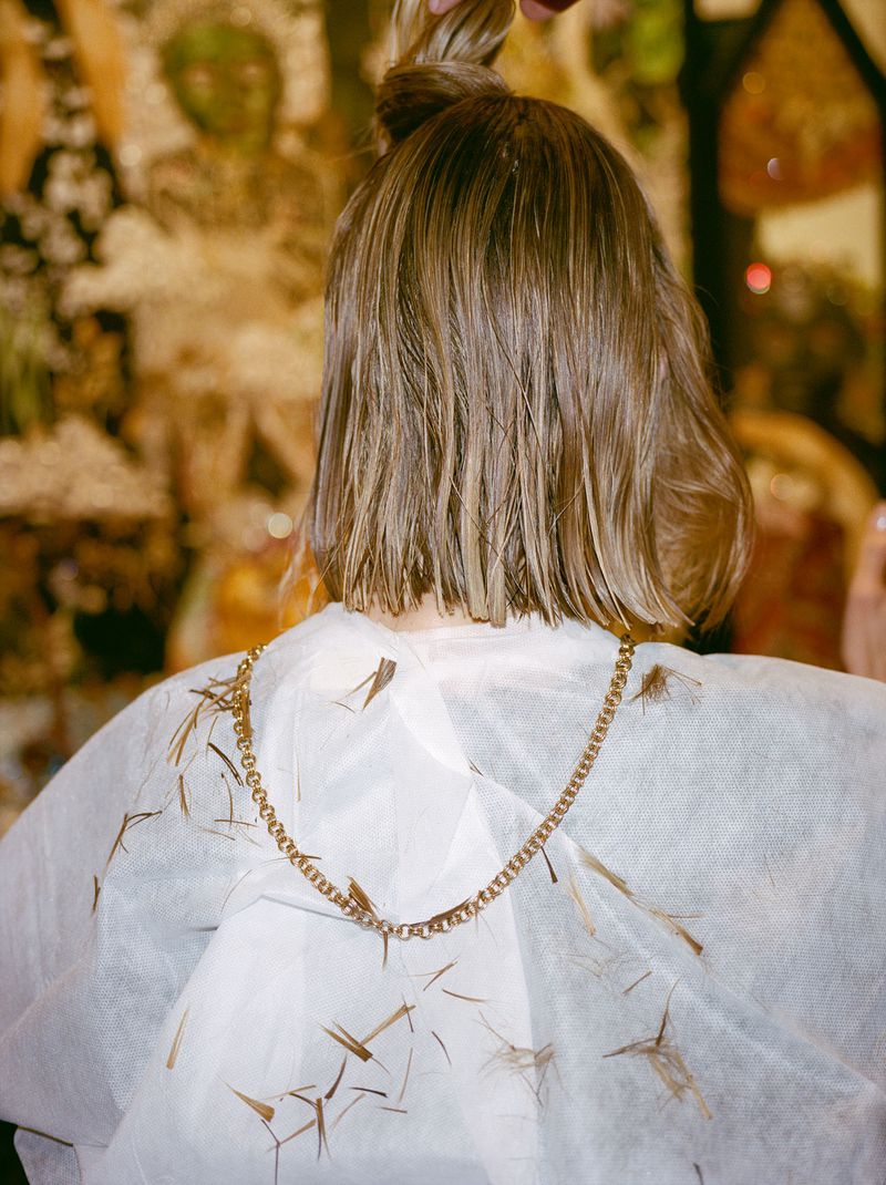 Back view of a blonde woman with a short bob haircut wearing a white cape covered in cut hair pieces and a gold chain necklace.