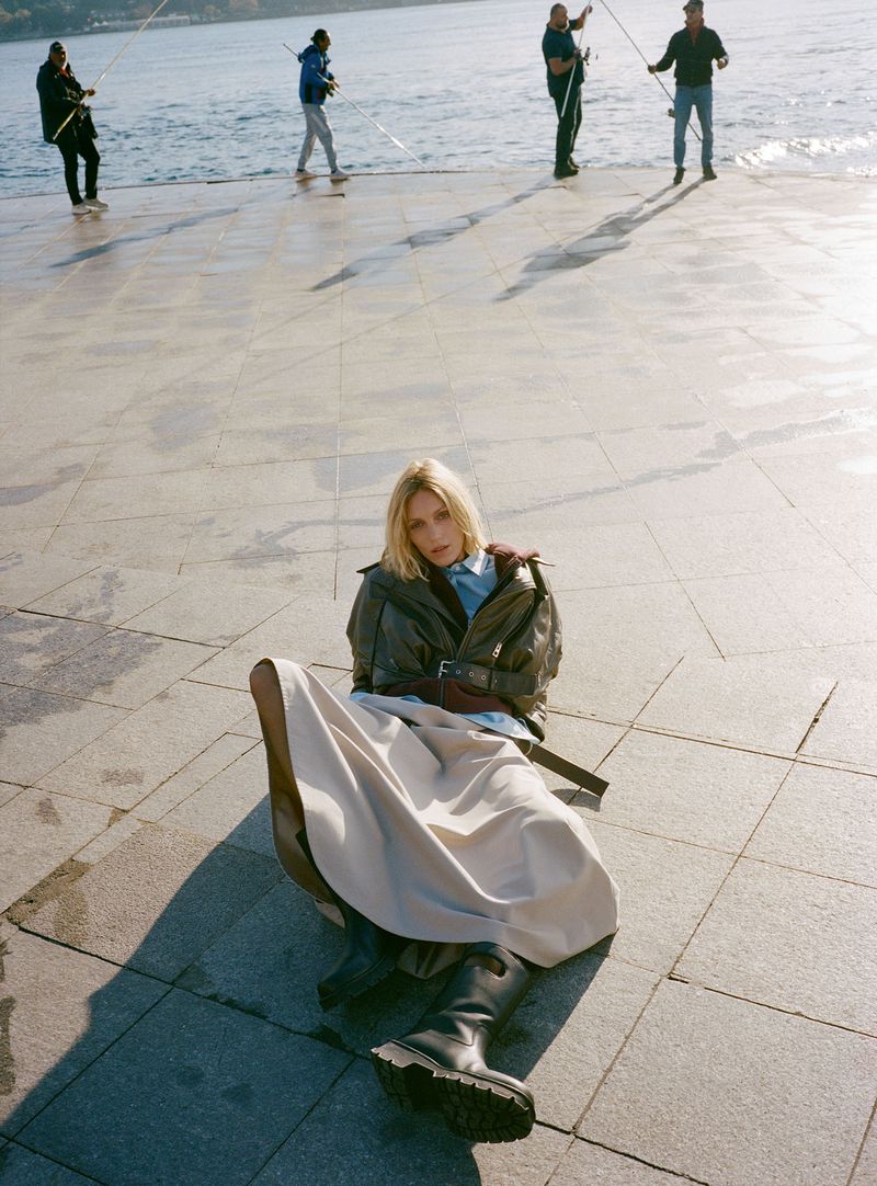 A blonde woman in a jacket and skirt is lying on a concrete platform; five men are visible in the distance fishing on the water's edge.