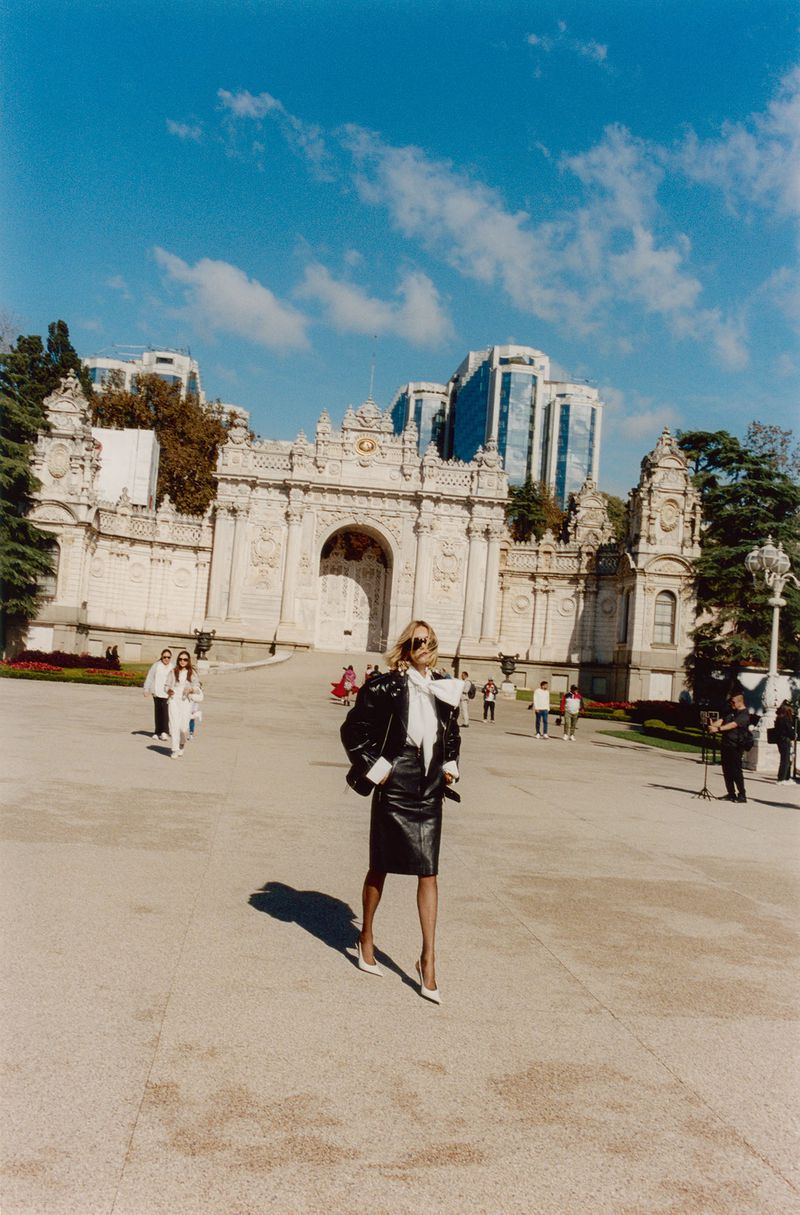 Woman in a black leather jacket and skirt walks confidently in front of an ornate archway and modern buildings on a sunny day.