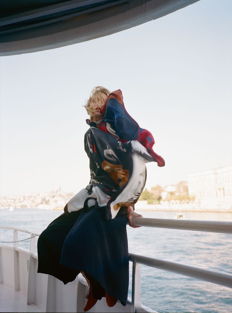 A blonde person perched on a metal railing on a boat deck, wrapped in a large, patterned silk scarf with flowing fabric against a river.