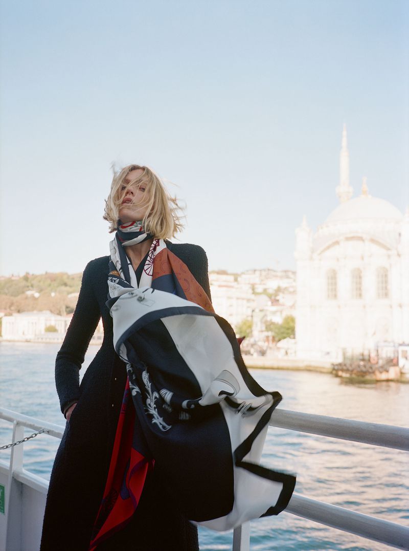 Woman in stylish scarf stands on a boat, with a white building in the background, under a clear sky, fashion photography.