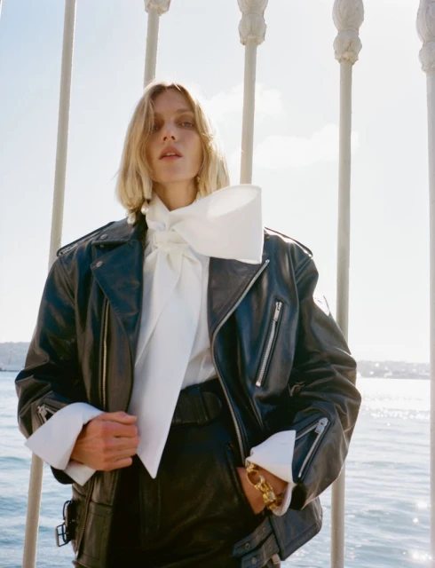 A blonde female model poses in a black leather jacket and white bow blouse on an outdoor terrace with light blue water behind the fencing.