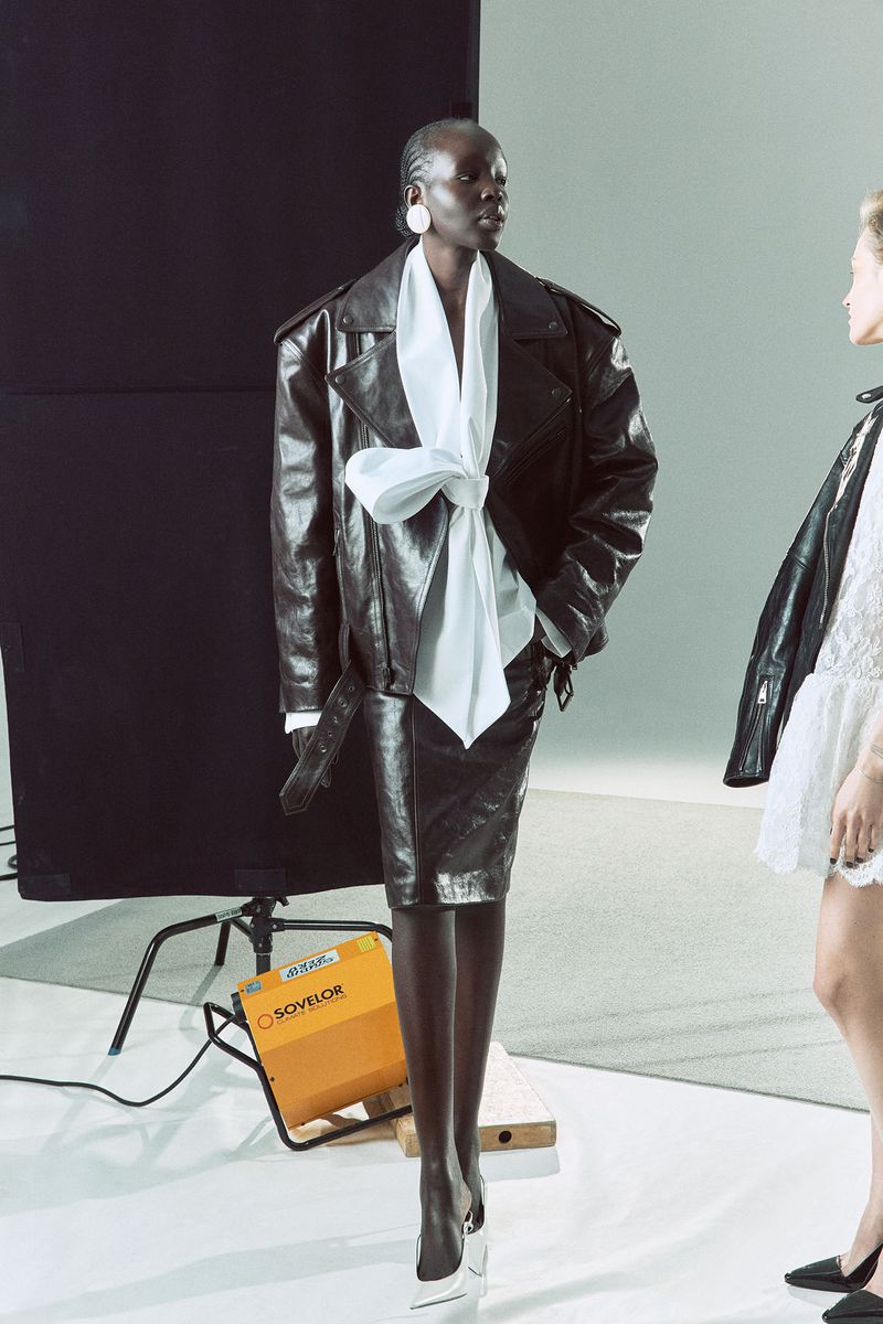 A runway shot of a model wearing a black leather jacket with a white shirt, black skirt and white heels in a studio setting.