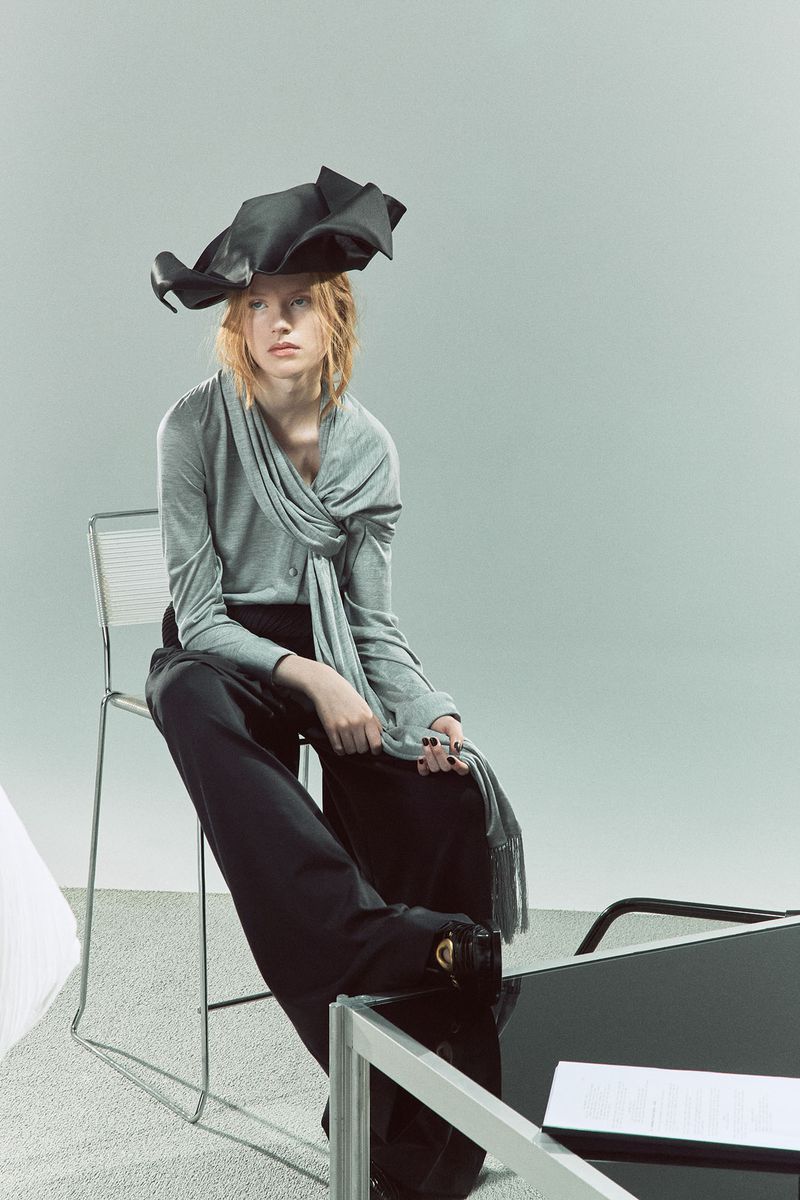 Model wearing a structured black sculptural hat, draped grey top, and black trousers while seated on a metal stool in a studio.
