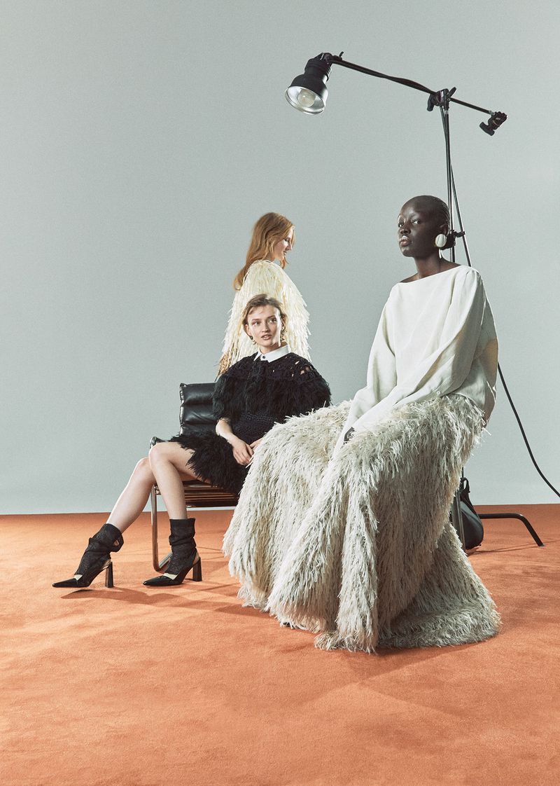 Three fashion models pose in a studio with a light stand. The models wear feathered dresses. Two are seated and one stands in the background.