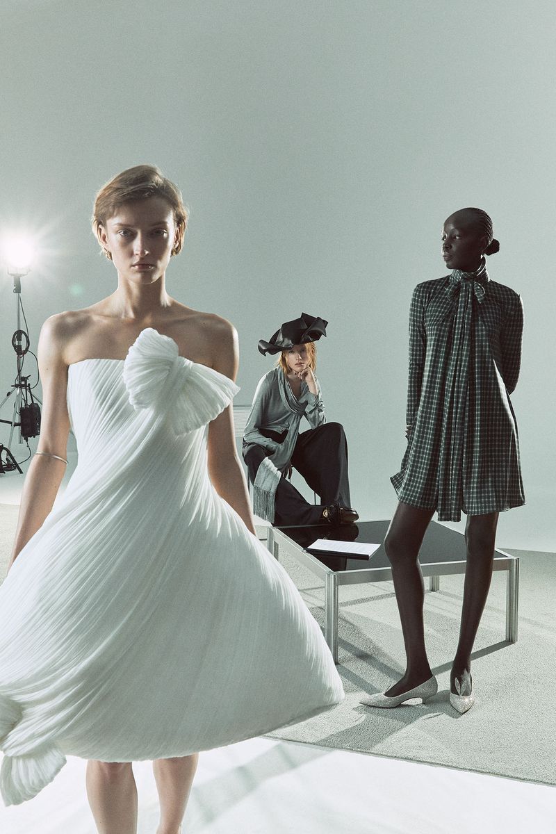 Three fashion models pose in an indoor photo studio. One wears a white strapless gown, one sits in dark pants, and one stands in a patterned dress.