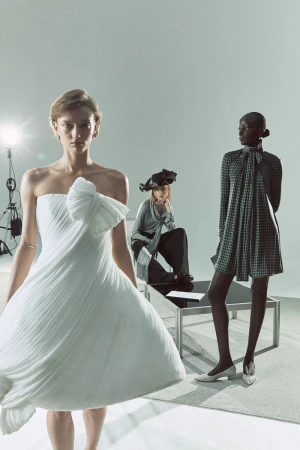 Three fashion models pose in an indoor photo studio. One wears a white strapless gown, one sits in dark pants, and one stands in a patterned dress.