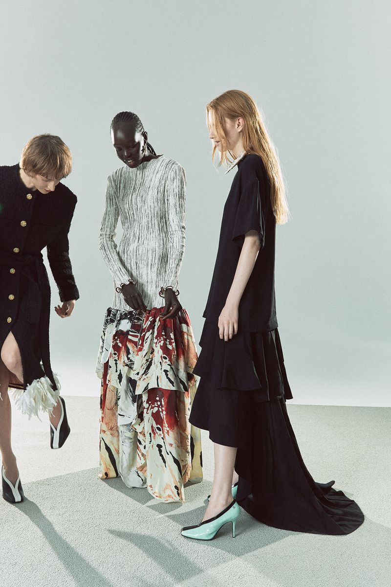 Three women in a studio wearing dresses, the woman on the left wearing a black dress, the woman in the center wearing a white top and printed skirt.