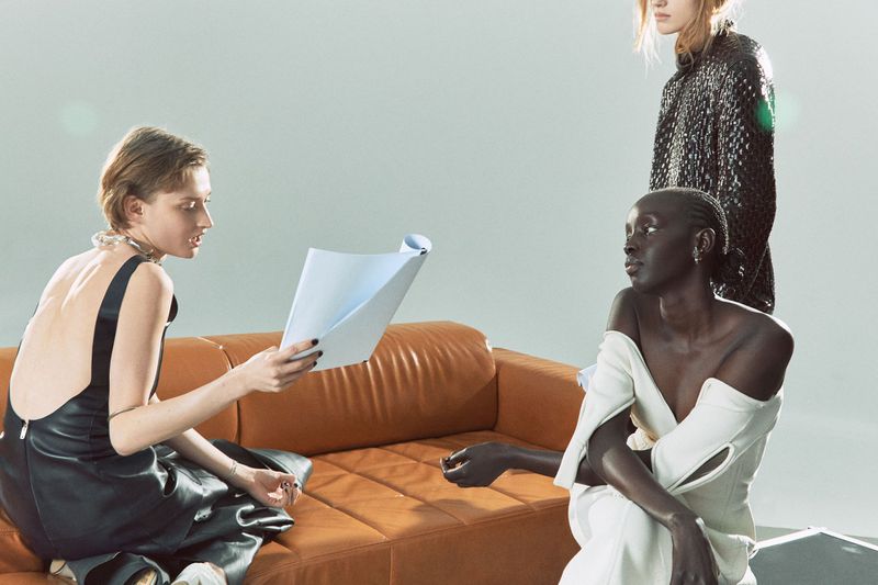 Three fashion models in a studio setting; one seated on a tan couch holds a white document, while two stand nearby in unique dresses.