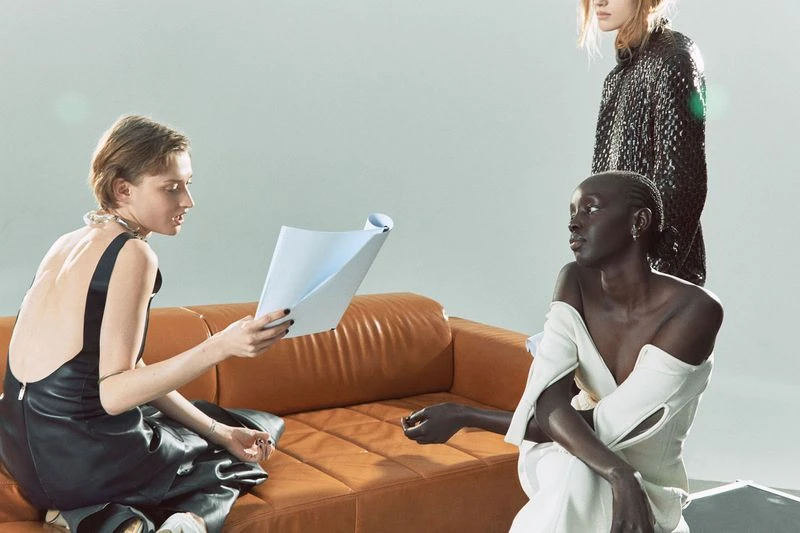 Three fashion models in a studio setting; one seated on a tan couch holds a white document, while two stand nearby in unique dresses.
