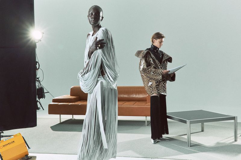 Fashion shot in a studio with two women wearing avant-garde outfits next to a tan leather couch and lighting equipment.