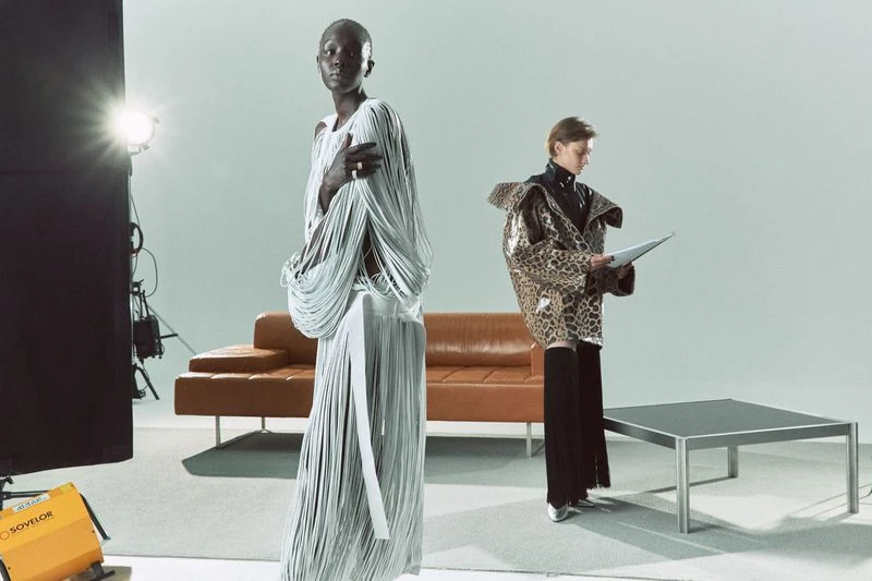 Fashion shot in a studio with two women wearing avant-garde outfits next to a tan leather couch and lighting equipment.
