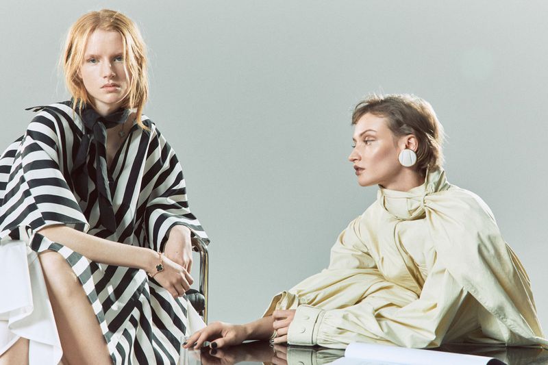 Two fashion models pose indoors in bright studio light. One wears black and white stripes and the other is in an off-white dress.