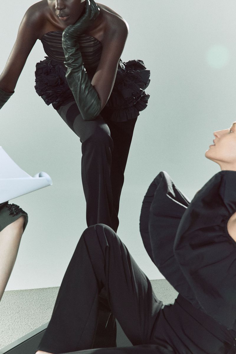 Two women model dramatic dark couture with angular poses and runway-ready looks against a light backdrop, showcasing fashion-forward design.