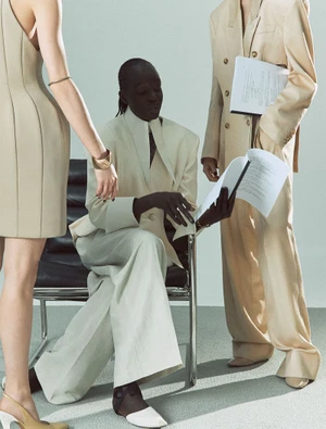 Akuol Deng Atem seated reading a script in an oversized beige suit between two models in tailored neutrals.