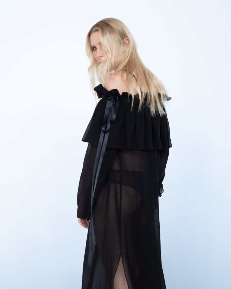 Blonde woman with shoulder-length hair looks back over her shoulder wearing a black, off-the-shoulder, sheer dress with ribbons.