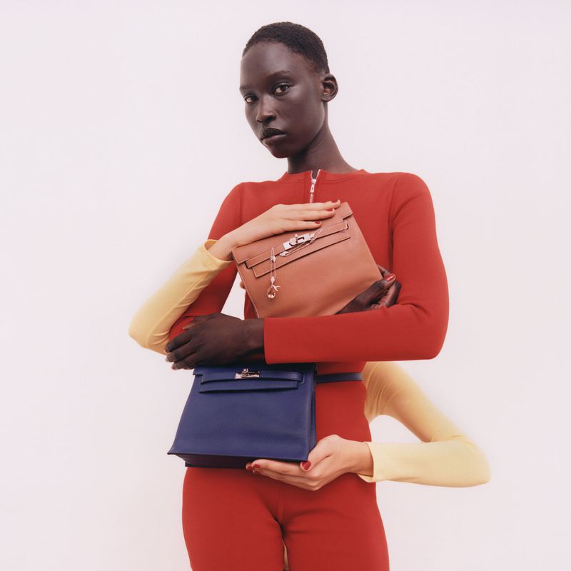 Fashion shot features a dark-skinned model holding two leather handbags in front of a white backdrop. The model wears a red catsuit.