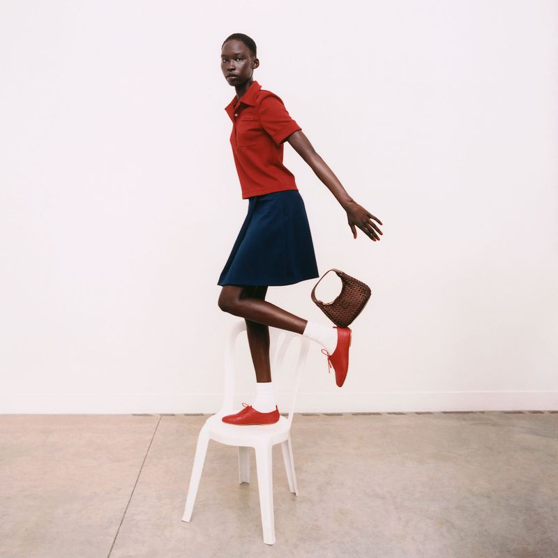 Fashion shot of a Black woman balancing on a white chair, wearing a red top, blue skirt, and carrying a woven handbag in a minimalist setting.