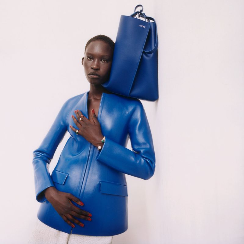 Fashion shot of Black woman wearing a blue jacket and carrying a blue bag. She poses against a white wall with studio lighting.