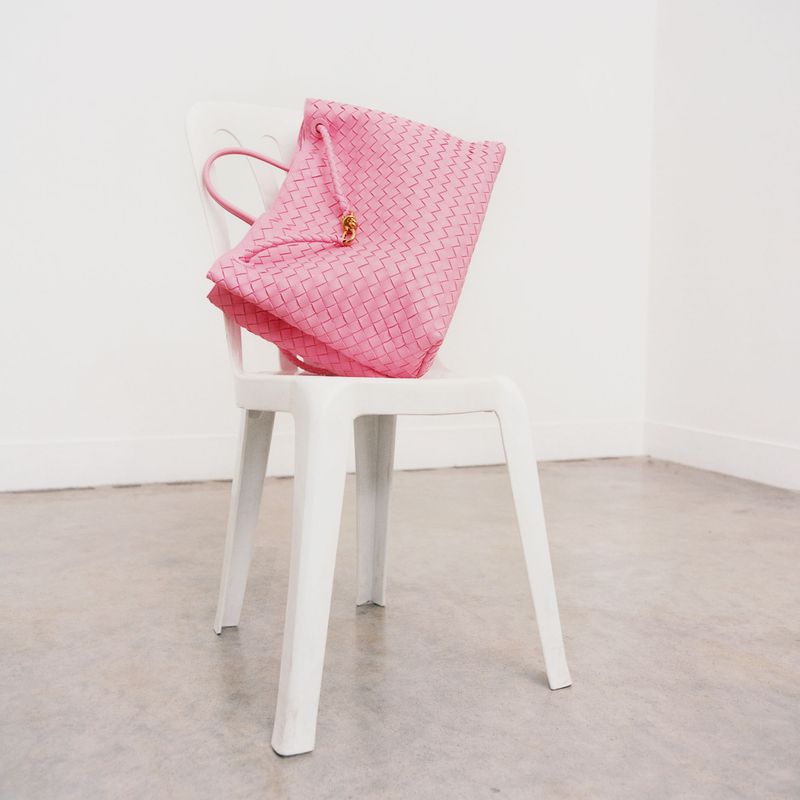Pink woven Bottega Veneta bag sits on a white plastic chair in an indoor setting with white walls and concrete floors.
