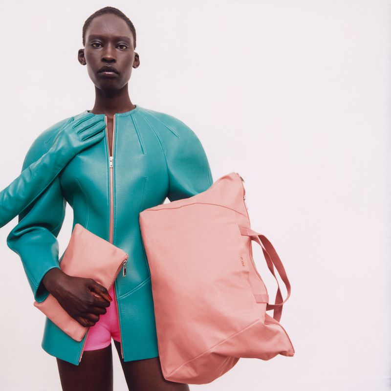 Editorial fashion shot of a Black female model in a teal jacket, gloves, and pink shorts, holding a light pink handbag and clutch against a white backdrop.