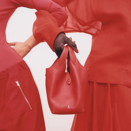 Close-up studio shot of a red leather handbag held by a person with dark skin tone, with a model partially visible in the background.