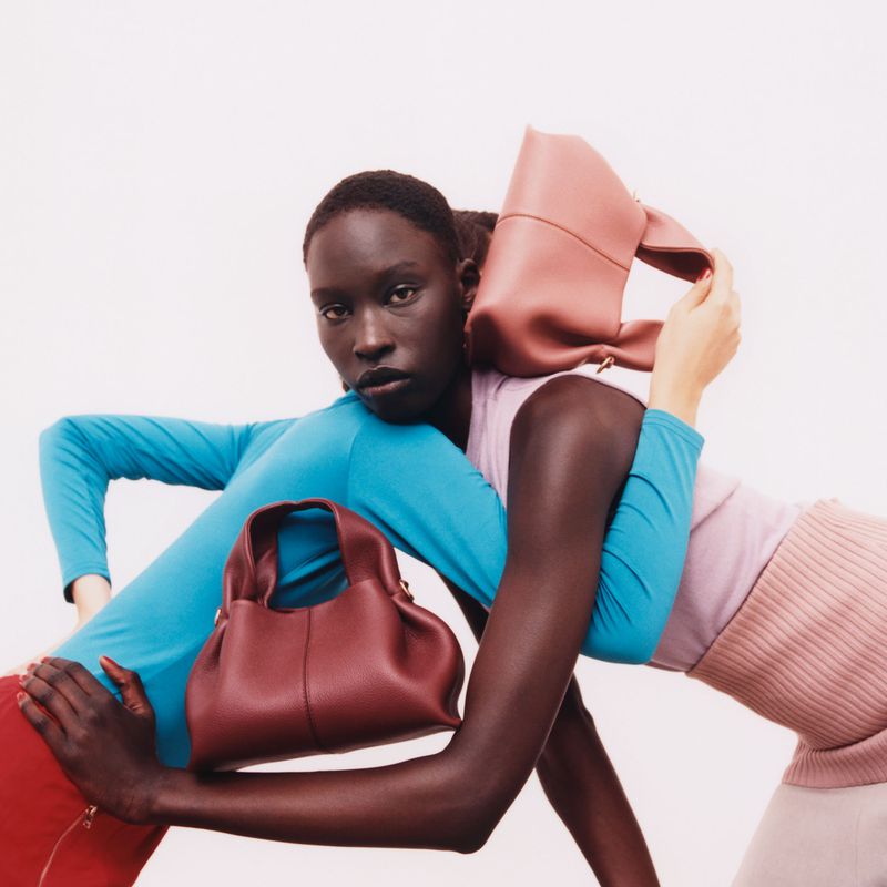 Two adult women pose with leather handbags against a white background in a fashion editorial shot with bright, studio lighting.