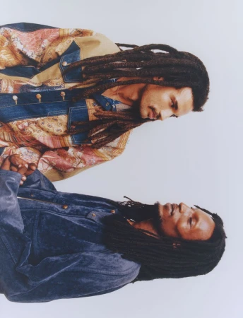 Two adult males with dreadlocks wearing denim and patterned jackets pose in a photo studio with a bright, neutral background.