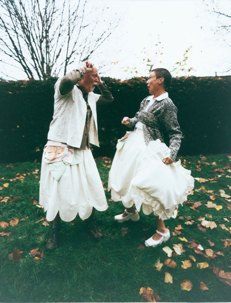 Two people in dresses pose outdoors in a green space with trees in the background, one person gesturing with hands to head, the other lunging forward.