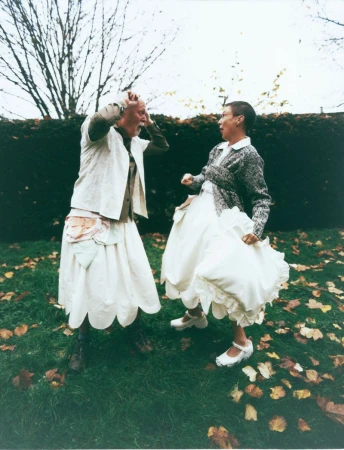 Two people in dresses pose outdoors in a green space with trees in the background, one person gesturing with hands to head, the other lunging forward.