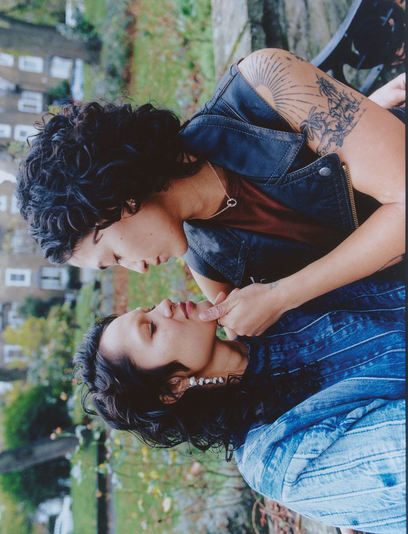 Two women in a tender moment outdoors, one resting on a bench while the other gently touches her face, buildings visible in the background.