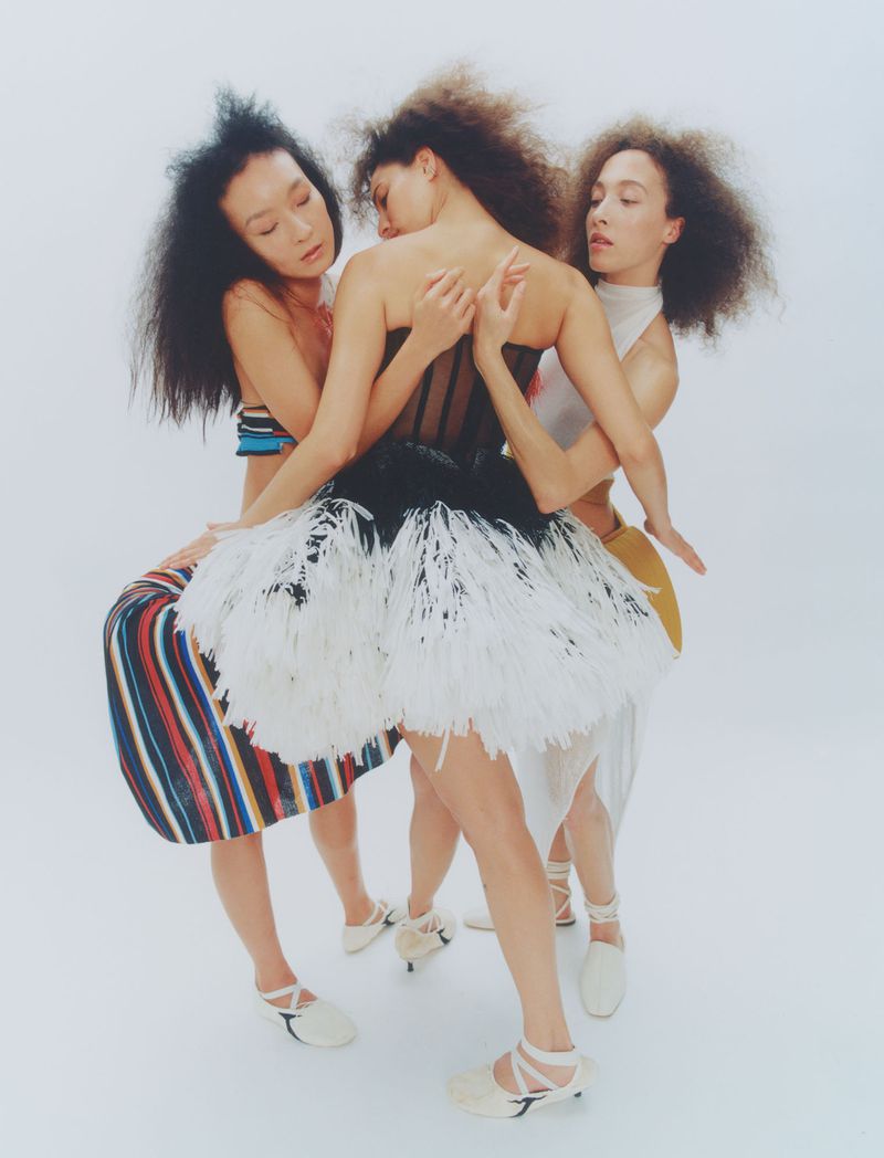 Three women in fashionable dresses pose in a bright studio. One model wears a tutu dress, and another a striped skirt with a cropped top.